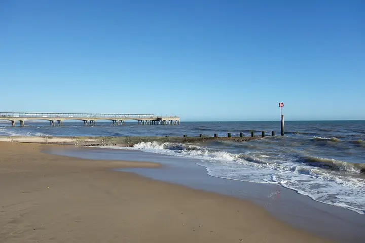 A sandy beach with gentle waves, a pier in the background, and a clear blue sky above.